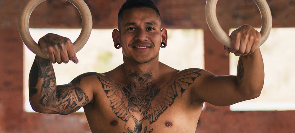 A topless man smiling at the camera in a gym setting, with stretched ear tunnels.