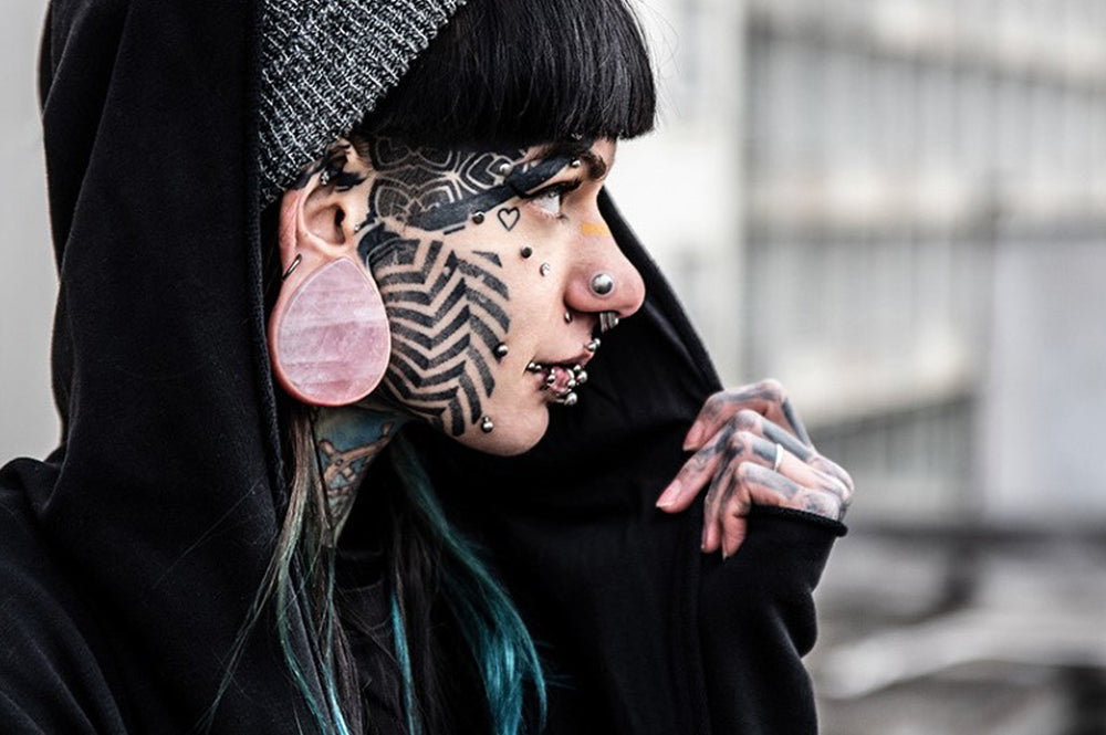 A women posing with face tattoos and multiple silver face piercings, showing her large stretched ear that features a pink stone teardrop ear plug
